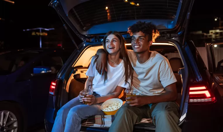 Diverse young couple having romantic date. Cheerful guy and his girlfriend watching a movie, sitting together in car trunk in front of a screen in an open air cinema
