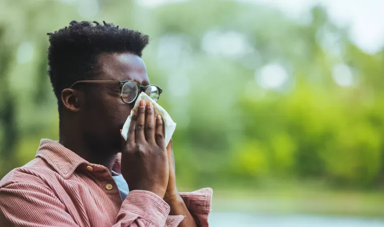 Allergic black man blowing on wipe in a park on spring season. Man with allergy or cold, blowing his nose with a tissue, looking miserable unwell very sick, isolated outside green trees background