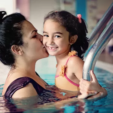 A mom in the pool giving her daughter a kiss on the cheek.