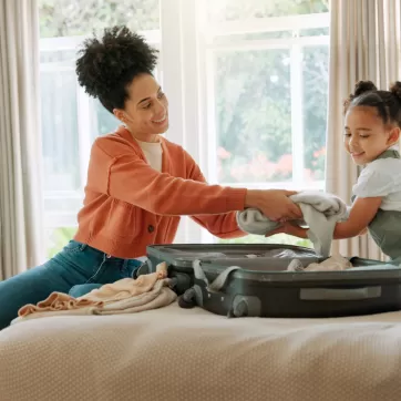 Travel, luggage and mom packing with child in bedroom getting ready for trip. Helping hands, black family and young girl help mother pack clothes in suitcase for holiday, vacation and weekend
