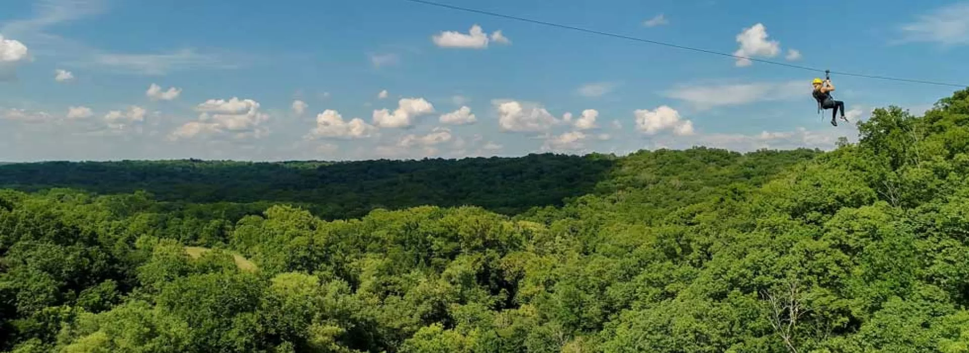 Drone shot of camper on zipline at Camp Kern's Ozone Ziplines on a sunny day with green trees and blue sky in the background